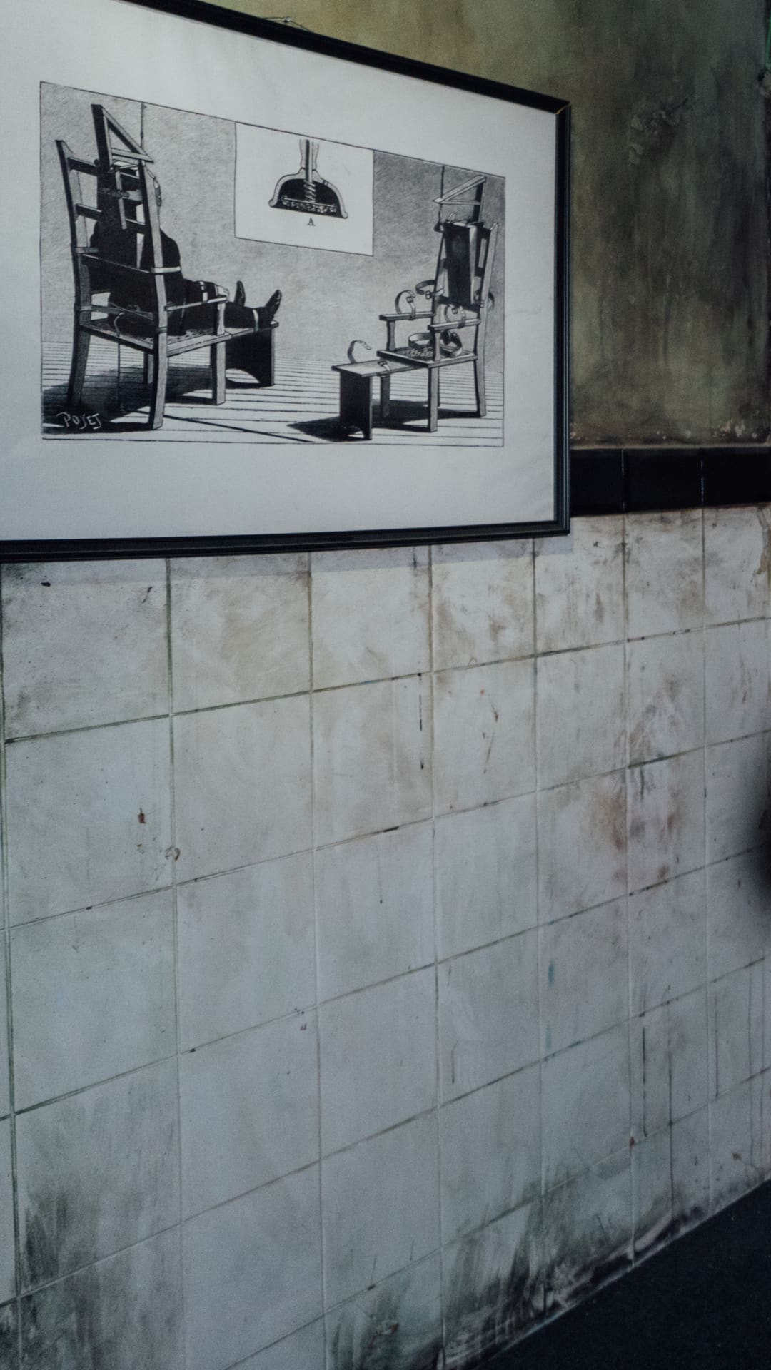 Laughing visitor taking a selfie beside a bloodied figure strapped into an electric chair with a metal head-cap, under a framed illustration of an antique execution chair at Museum of Madness Barcelona