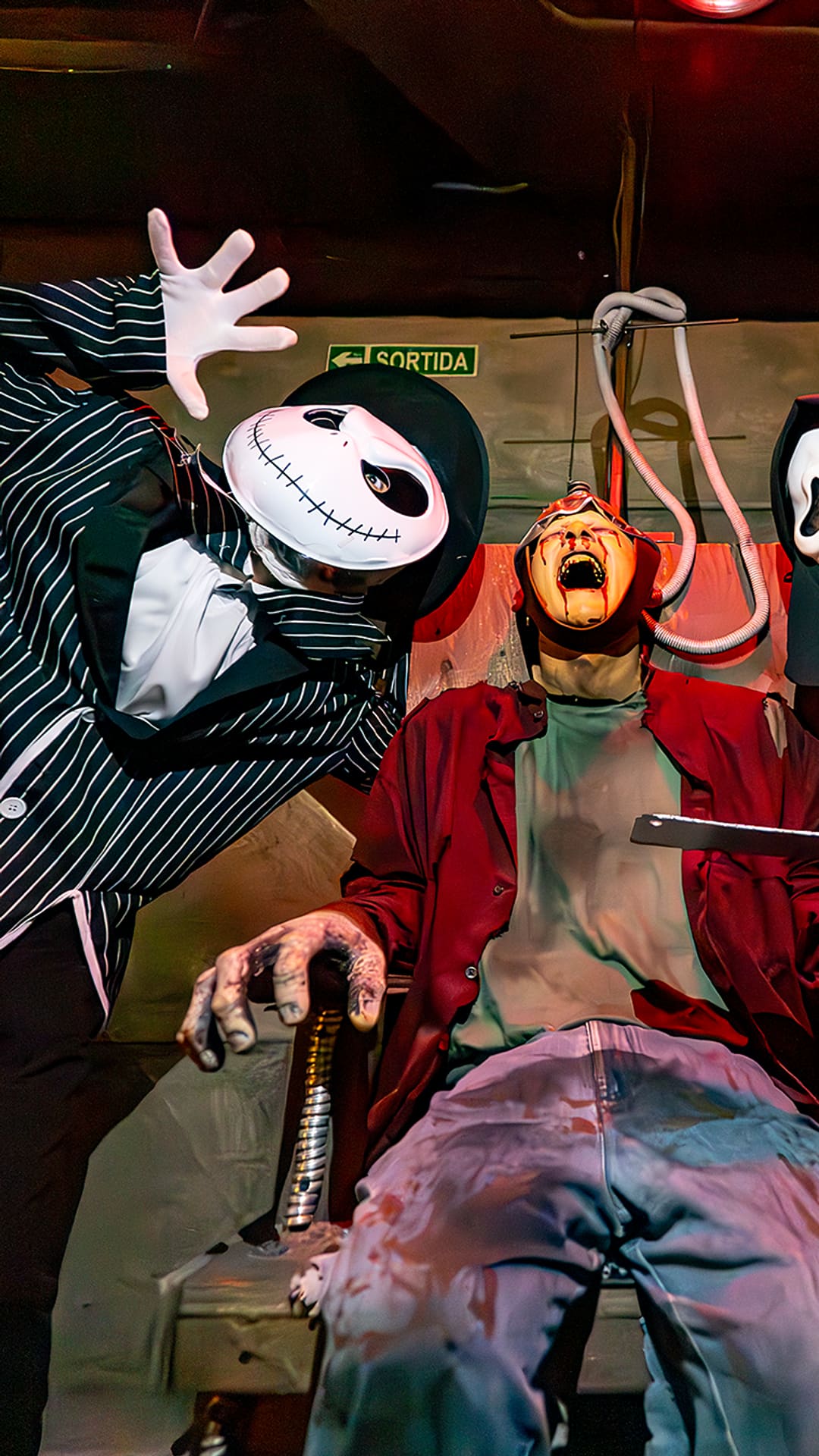 Three masked figures looming over a screaming victim strapped to a chair, one holding a straight razor, under harsh red light at Museum of Madness Barcelona
