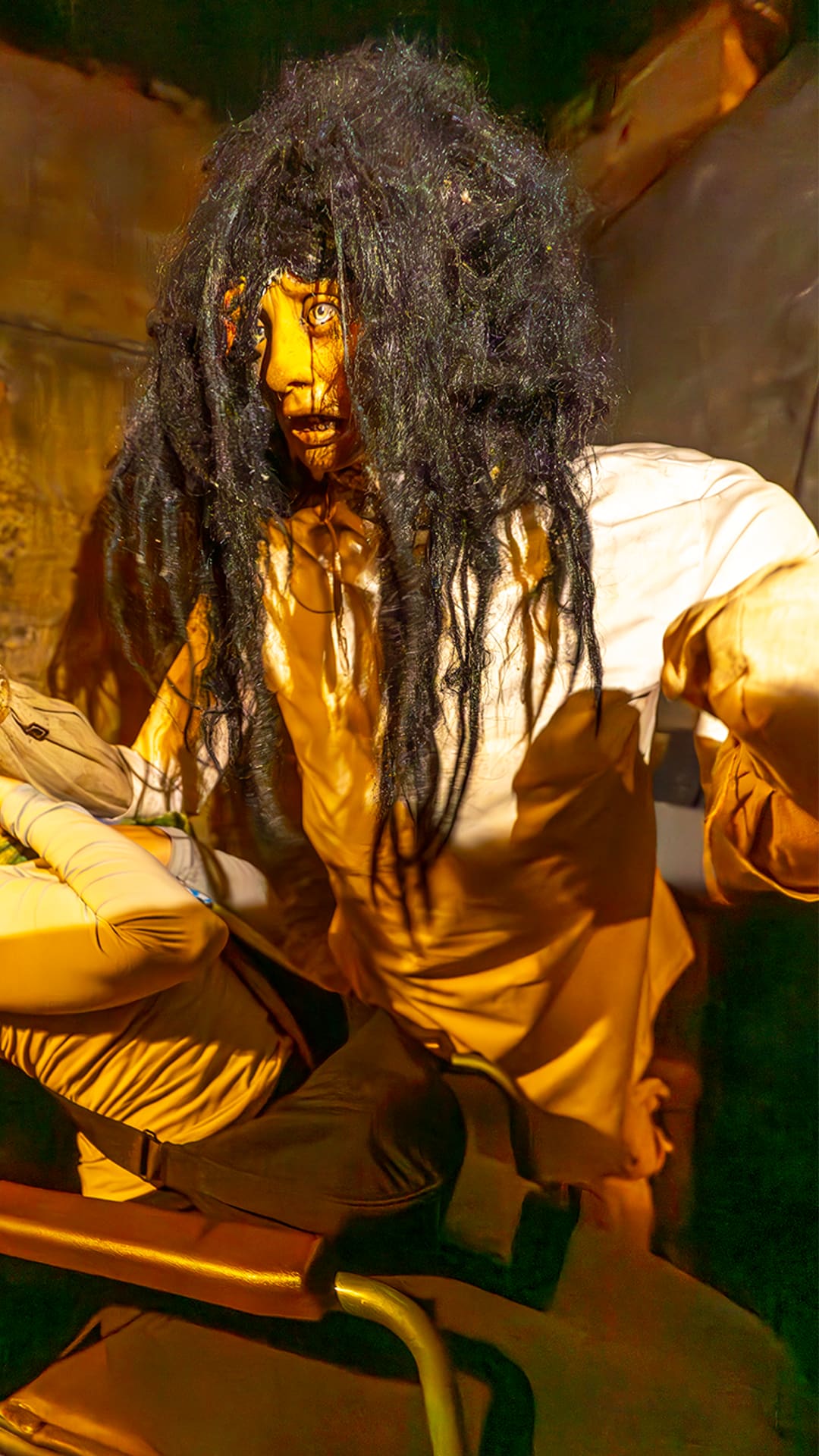 A wild-haired 'patient' figure in a stained gown lunging at a laughing visitor in a wheelchair, under amber light at Museum of Madness Barcelona